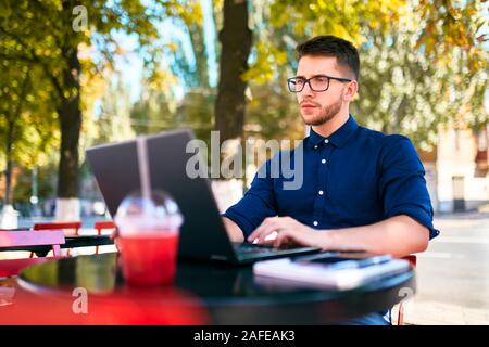 Young attractive hipster freelancer working remotely on laptop at outdoor cafe. Businessman browsing internet at coffeshop. Blogger posts to social Stock Photo