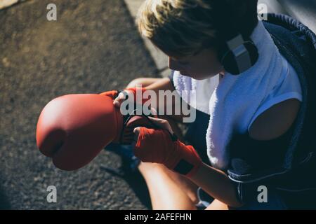 Handsome young male with boxing bands in his hands, looking gloves sitting on the road. Boy prepares for a boxe workout, listening music. Sport, youth Stock Photo