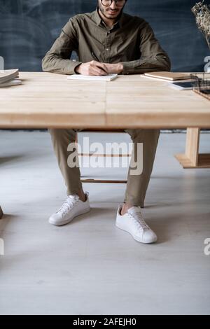 Young casual man sitting by wooden table in office or classroom and making notes Stock Photo