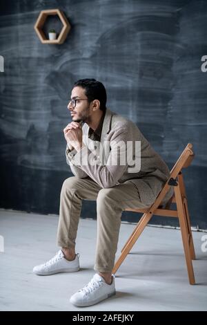 Young pensive businessman in smart casual sitting on chair in front of camera Stock Photo