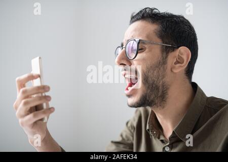 Young bearded man in eyeglasses and shirt shouting in front of smartphone camera Stock Photo