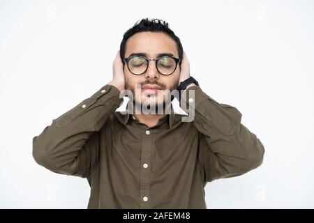 Young annoyed man in eyeglasses keeping his eyes closed and hands on ears Stock Photo