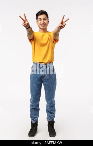 Vertical full-length shot cheerful hipster asian guy in jeans, yellow t-shirt, extend arms forward showing peace or victory signs and smiling, express Stock Photo