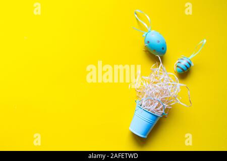 Decorative colored eggs and a straw nest in a miniature bucket on a bright yellow background, minimalistic easter background or concept Stock Photo