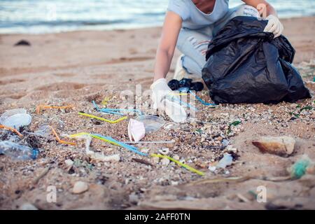 Woman in blue shirt with white gloves and big black package collecting garbage on the beach. Environmental protection and planet pollution concept