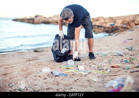 Man in dark shirt and shorts with white gloves and big black package collecting garbage on the beach. Environmental protection and planet pollution co Stock Photo