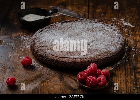 Chocolate mud cake, swedish kladdkaka or sticky chocolate cake with powdered sugar. The cake is on a rustic, vintage wooden table with raspberries and