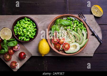 A fresh, healthy salad with zoodles zucchini noodles, baby tomatoes, avocado and edamame beans. The salad is in a wooden bowl on a dark bamboo table o Stock Photo