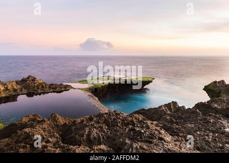 Devil's Tear at sunset, island of Nusa Lembongan, Bali, Indonesia. Rocky shore in foreground. Yellow sky with clouds beyond. Stock Photo
