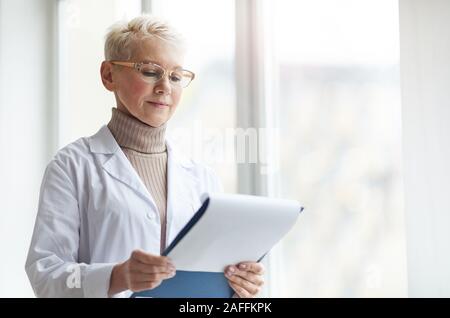 Waist up portrait of mature female doctor holding clipboard while standing by window in clinic and reading patient forms, copy space Stock Photo