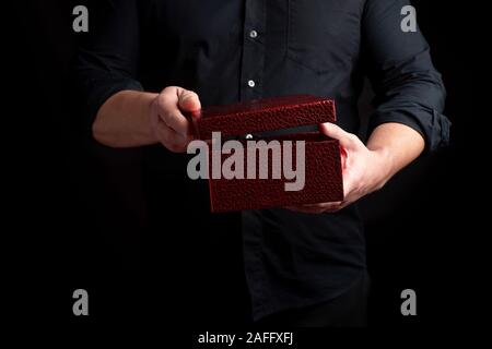 adult man in a black shirt holds a cardboard red gift box and opens the lid, dark background Stock Photo