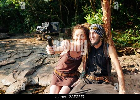 Happy mature couple sitting on stone in forest and taking selfie on smartphone attached to monopod Stock Photo
