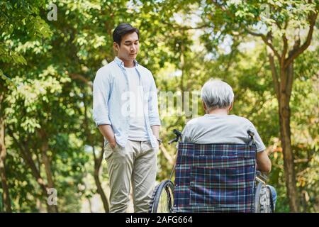 asian son talking to sad and depressed wheelchair bound father Stock Photo