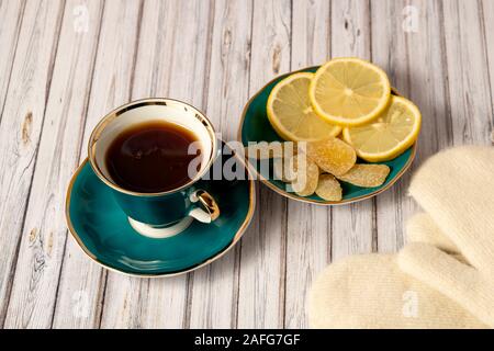 Green cup of hot tea, saucer with lemons and candied ginger and white mittens on a wooden table Stock Photo