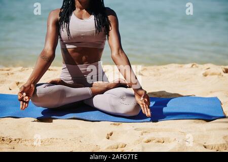 Cropped image of young Black woman in sports leggings sitting on beach in lotus position and enjoying meditation process Stock Photo