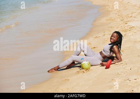 Happy young fit Black woman resting on beach with coconut cocktail after jogging in the morning Stock Photo