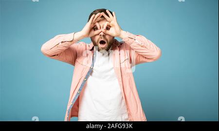 Strange bearded man makes a gesture like binoculars, pressing his hands to face, eyes look through fingers and is surprised. Stock Photo