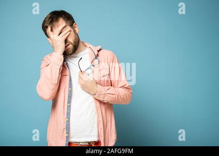 Tired or upset bearded man covers face with palm, holding glasses in hands. Blue background. Stock Photo