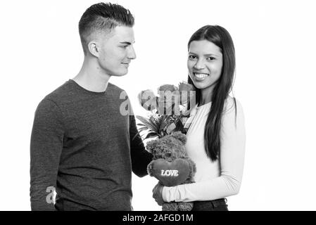 Young happy couple smiling and in love with woman holding red roses and teddy bear with heart and love sign Stock Photo