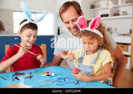 Father With Children Wearing Rabbit Ears Decorating Easter Eggs At Home Together Stock Photo