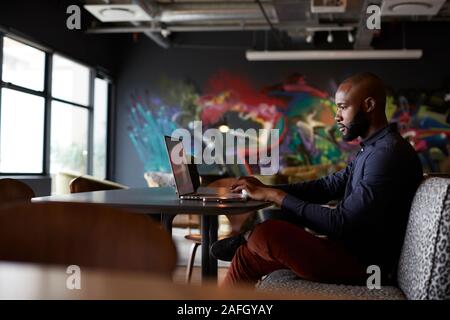 Mid adult black male creative sitting at table in office dining area using a laptop, side view Stock Photo