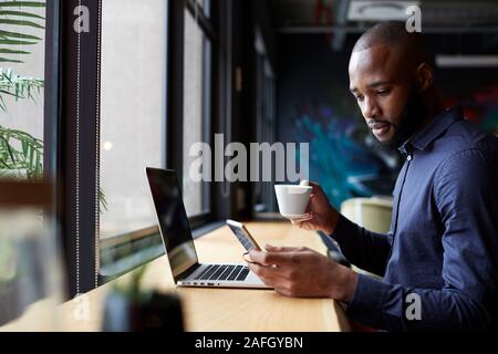 Mid adult black male creative sits by window having coffee, using a laptop and smartphone, side view Stock Photo