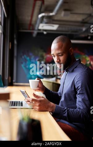 Mid adult black male creative sitting by window having coffee using smartphone, side view, vertical Stock Photo