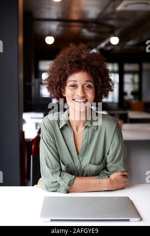 Millennial black female creative sitting at desk in an open plan office smiling to camera, vertical Stock Photo