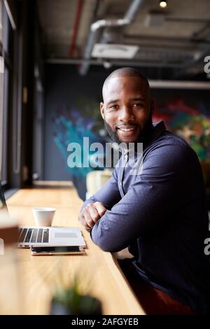 Black male creative sitting by window in cafe using laptop, turning and smiling to camera, vertical Stock Photo