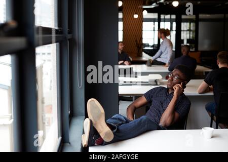 Young black male creative sitting in an office with his feet up on the desk using phone, close up