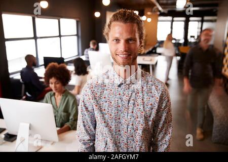 Millennial white male creative standing in a busy casual office, smiling to camera Stock Photo
