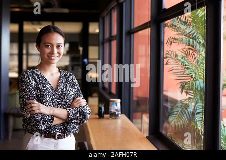 Millennial Asian businesswoman standing by the window in an office smiling to camera, waist up Stock Photo