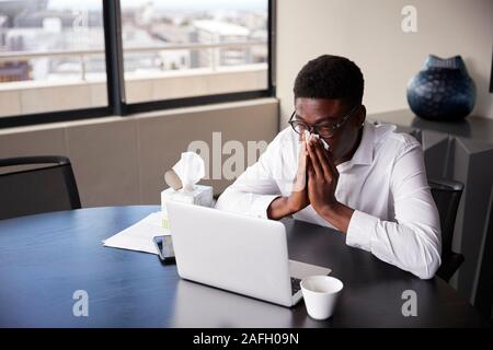 Young black businessman sitting at an office desk blowing his nose into a tissue, elevated view Stock Photo
