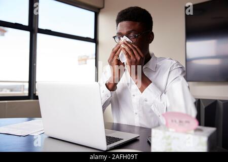 Young black businessman sitting at an office desk blowing his nose into a tissue Stock Photo