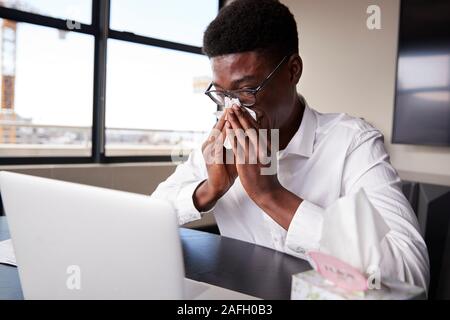 Young black businessman sitting at an office desk blowing his nose into a tissue, close up Stock Photo