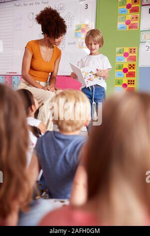 Male Pupil In Elementary School Classroom Reading Book To Class With Teacher Stock Photo