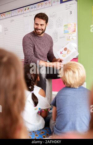 Male Teacher Reading Story To Group Of Elementary Pupils In School Classroom Stock Photo