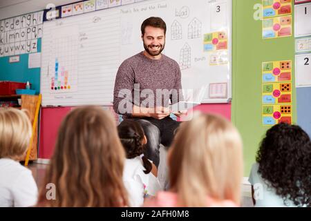 Male Teacher Reading Story To Group Of Elementary Pupils In School Classroom Stock Photo