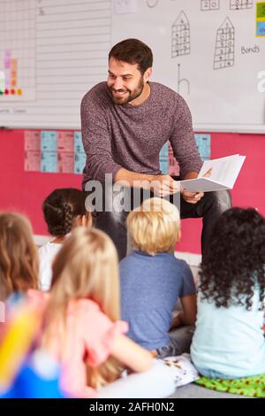 Male Teacher Reading Story To Group Of Elementary Pupils In School Classroom Stock Photo