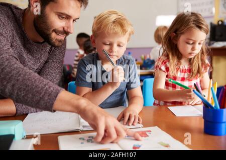 Elementary School Teacher Helping Pupils As They Work At Desk In Classroom Stock Photo