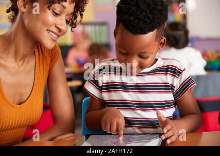 Elementary School Teacher And Male Pupil Drawing Using Digital Tablet In Classroom Stock Photo