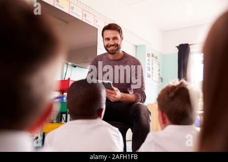Male Teacher Holding Digital Tablet Teaches Group Of Uniformed Elementary Pupils In School Classroom Stock Photo