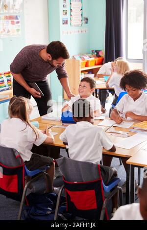 Male Teacher Holding Digital Tablet Teaches Group Of Uniformed Elementary Pupils In School Classroom Stock Photo