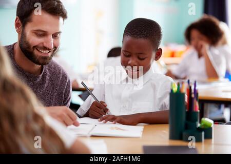 Elementary School Teacher Giving Male Pupil Wearing Uniform One To One Support In Classroom Stock Photo