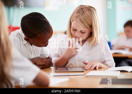 Two Elementary School Pupils Wearing Uniform Using Digital Tablet At Desk Stock Photo