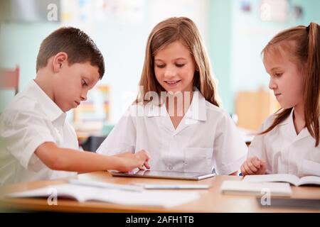 Three Elementary School Pupils Wearing Uniform Using Digital Tablet At Desk Stock Photo