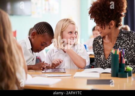 Female Teacher With Two Elementary School Pupils Wearing Uniform Using Digital Tablet At Desk Stock Photo