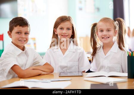 Portrait Of Three Elementary School Pupils Wearing Uniform Using Digital Tablet At Desk Stock Photo