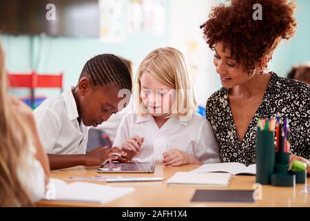 Female Teacher With Two Elementary School Pupils Wearing Uniform Using Digital Tablet At Desk Stock Photo