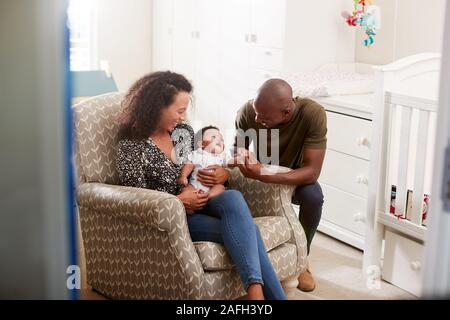 Loving Parents Sitting In Chair Cuddling Baby Son In Nursery At Home Stock Photo